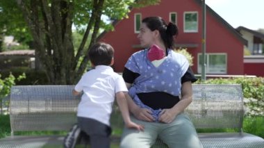 Anne yeni doğmuş bebeğini sarmalayıp bankta oturuyor ve yanında küçük bir çocuk var. Yeşillik ve ikamet geçmişi olan bir parkta hassas bir aile anını yakalıyor.