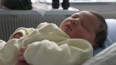 Yeni doğmuş bebek annesinin kollarında huzur içinde dinleniyor, rahat bir krem kıyafet giymiş. Yakın çekim sakin bir ifade ve sevgi dolu bir kucaklama, bebekle ilk zamanların sıcaklığını ve sakinliğini vurguluyor. 