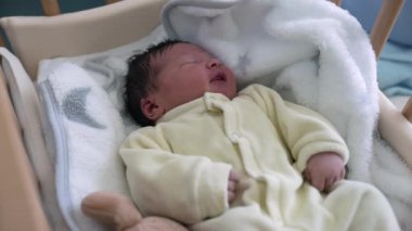 Bebek beşikte huzur içinde dinleniyor, yumuşak beyaz bir battaniyeye sarılıyor, erken çocukluğun huzurlu ve rahat atmosferini yakalıyor ve hayatın ilk günlerinde sunulan şefkatli bakımı alıyor.