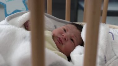 Bebek beşikte uyandı, yumuşak bir battaniyeye sarıldı, etrafı gözetliyor. Merak ve masumiyet, rahat bir ortamın rahatlığını ve sıcaklığını vurguluyor.