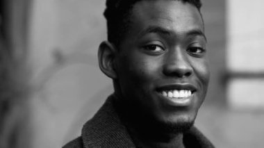 Siyah ceket ve balıkçı yaka giyen gülümseyen Afro-Amerikan genç adam. Neşeli ve kendinden emin bir ifadeye sahip, bulanık şehir geçmişli siyah beyaz bir portre.