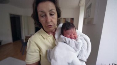Büyükannem yeni doğmuş bir bebeği sıcak beyaz bir battaniyeye sarmış, ev ortamında duruyor. Bebek biraz huysuz, ve büyükanne endişeli görünüyor, bir anlık ilgi ve ilgi gösteriyor.
