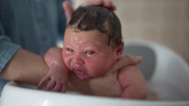 Yeni doğan bebek banyo yapıyor, ebeveyni tarafından nazikçe tutuluyor, bebeğin derisine su damlacıkları dökülüyor, gözleri açık, ilk yaşam deneyimleri, bakım ve bakım, sıcak, samimi bir ortamda.