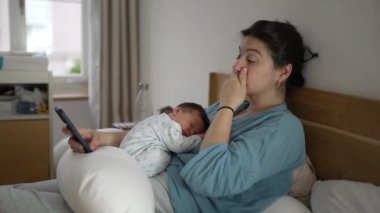 Yeni doğmuş bir bebeğin göğsünde uyumasından yorgun bir anne, akıllı bir telefon tutarken gözlerini ovuşturması, rahat kıyafetler giymesi, ev ortamında samimi olması, yorgunluk ve sevgiyi göstermesi
