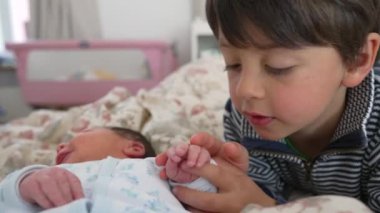 Yeni doğan bebeğin minik elini tutan küçük çocuk, kapalı yatak odası ortamında, nazik etkileşim, kardeş bağı, sevgi ve koruma ifadeleri, aile anı, yeni başlangıçlar 