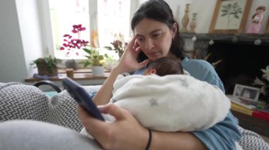 Anne, yeni doğmuş bebeğini kucağında tutarken, telefona bakarken, arka planda bitkiler ve dekoratif elementlerle dolu parlak bir odada koltukta oturuyor.