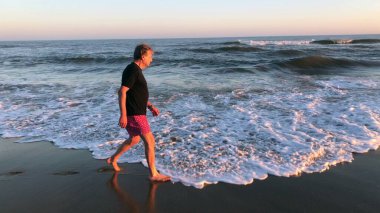 Older man walking at the beach during sunset, meditative senior person walks by the shore