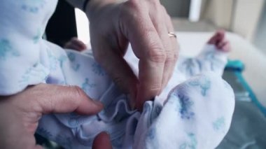 Ebeveynler, bebek kıyafetinin küçük bir düğmesini nazikçe açarak yeni doğmuş bir bebeği giydirmenin detaylarını vurguluyor. Eller ve elbiseler, aşkı ve korumayı simgeliyor.