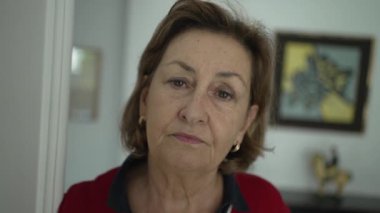 Sarı saçlı, yeşil gözlü, altın küpeli ve kırmızı bluzlu, arka plandaki resimlerle oturma odasında oturan ve kameraya ciddi ciddi bakan zarif olgun kadının yakın çekimi.