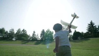 Sinematik Çekim - Kamera, Güneş Işığına Karşı Koşan Çocuğu Elinde Planör Modeli ile Yeşil Park Bölgesinde Uzaktan Uçuşla İzliyor.