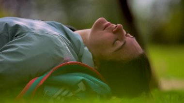 Kapalı Gözlerle Rahatlayan Kadın 'ın Yakın Çekimi, Gündüz vakti Yaprak Park' ta Çimlere Uzanmak, Sırt çantasını Yastık Otu Olarak Kullanmak