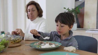 Genç çocuk büyükannesinin yardımıyla yemek masasında yemek yiyor. Aile birlikteliği anı, nesiller arasındaki beslenme bağını gösteriyor.