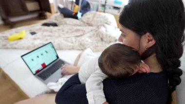 Anne kucağında yeni doğmuş bir bebek varken dizüstü bilgisayarda çalışıyor, mesleki görevlerin ve ebeveynlik sorumluluklarının dengesini gösteriyor, modern aile yaşamını ve çoklu görev becerilerini vurguluyor