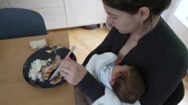 Anne tek eliyle yemek yiyor, kucağında uyuyan yeni doğmuş bir bebek, yeni anneliğin günlük dengesini ve anne ile çocuk arasındaki bozulmaz bağı vurguluyor.