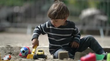 Etrafı oyuncaklarla çevrili olan çocuk, Kum Çukuru Kazılarında Küçük Plastik Kürek kullanarak Sunny Skies 'in altında açık havada çukurlar kazıyor.