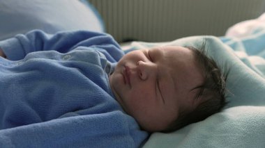 Yeni doğmuş bir bebeğin, yumuşak bir battaniyenin üzerinde mavi tulumla huzur içinde uyuduğu yakın plan. Yeni doğmuş bir bebeğin rahat ve sakin bir ortamda uykusunun narin ve huzurlu doğasını yakalamak.