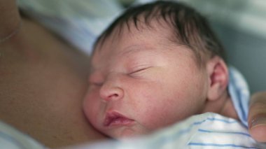 Annenin göğsüne yaslanmış mavi beyaz çizgili bir gömleğe sarılı yeni doğmuş bir bebeğin yakın çekimi. Anne tarafından cilt teması yoluyla sağlanan sıcaklık ve güvenlik