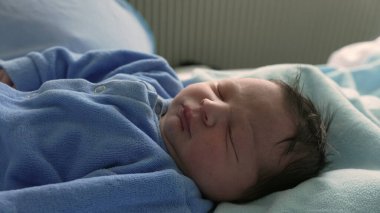 Yeni doğmuş bir bebeğin, yumuşak bir battaniyenin üzerinde mavi tulumla huzur içinde uyuduğu yakın plan. Yeni doğmuş bir bebeğin rahat ve sakin bir ortamda uykusunun narin ve huzurlu doğasını yakalamak.