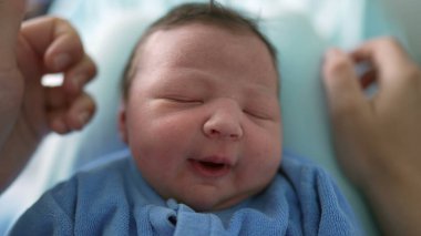 Yeni doğmuş bir bebeğin, yumuşak mavi bir tuluma sarılı, mışıl mışıl uyuduğu yakın plan. Annenin eli, bebeğin yüzünü okşar. Erken yaşların hassas ve hassas doğasını, sakin bir anda ön plana çıkarır.