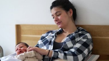 Anne yeni doğmuş bebeğe yatakta gülümsüyor, erken anneliğin neşesini ve bağını gösteriyor, anne ile çocuk arasındaki samimi sevgi ve bağ anını gösteriyor, aile hayatı bakımını ön plana çıkarıyor.