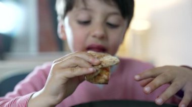 Bir çocuğun pizza diliminin tadını çıkarırken, yemekteki neşe ve nişanı gösterirken. Çocuğun yüzüne ve ellerine odaklanmak, yemekten zevk almanın duyusal deneyimini vurgular.