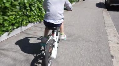 Kaldırımda bisiklete binen bir çocuğun geriye bakış açısı, genç sürücünün bağımsızlığını ve güvenini gösteriyor. Huzurlu bir mahalle ve bisiklet sürmenin sevinci
