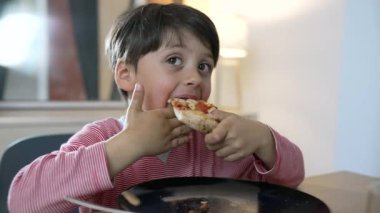 Çocuk yemek masasında bir dilim pizzanın tadını çıkarıyor, yemek sırasında çocukluğun neşesini ve masumiyetini yakalıyor. çocuğun içerik ifadesi ve yeme eylemi