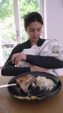 Anne çoklu görev, yeni doğmuş bebeğini sıcacık beyaz bir battaniyeye sarılı tutarken yemek yemek, huzurlu bir evde ebeveynliğin dengesini göstermek.