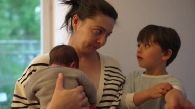 Anne, büyük çocuğunu azarlarken yeni doğmuş bebeğini bir inançsızlık ifadesiyle kucaklıyor. Aile ilişkilerinin dinamiği ve annenin özenli doğası.