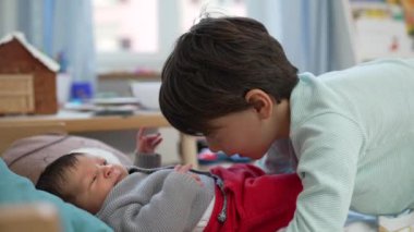 Büyük kardeş, sevgi ve sıcaklıkla dolu sıcak bir odada şefkatli ve doyurucu bir an yaratarak, bir yatakta uzanan bebeğe sevgiyle bakar.