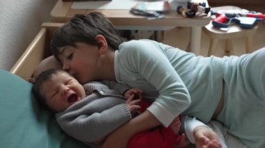 Büyük kardeş yeni doğmuş bir bebeğin yanında yatıyor. Sevgi ve bağlılık dolu bir anı paylaşıyor. Sıcak bir yuva ortamında kardeşler arasındaki sevgi ve bağlantıyı gösteriyor.