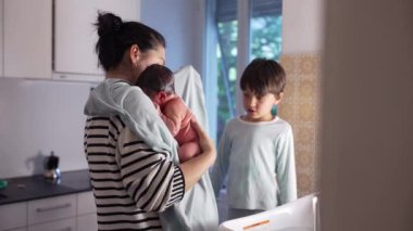 Anne, yeni doğmuş bebeğini banyodan sonra dikkatlice giydiriyor, küçük kardeşi evde hassas bir aile anını yakalıyor.