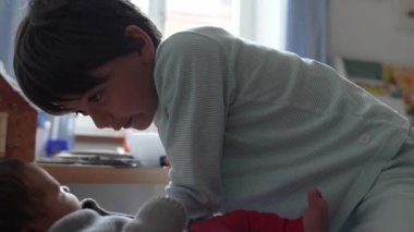 Büyük kardeş ve bebek bir yatakta sevgi dolu, güneşli, oyuncaklarla dolu sıcak bir odada hassas ve neşeli anlar paylaşıyorlar, güçlü bir kardeş bağı ve neşeli bir aile ortamı sergiliyorlar.