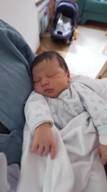 Anne, kucağında huzur içinde uyuyan yeni doğmuş bebeğine sevgiyle bakıyor. Sıcak beyaz bir battaniyeye sarılıp bir yuva ortamında annelik bağı ve şefkati vurguluyor.