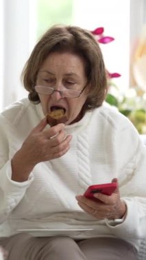 Gözlüklü yaşlı bir kadın, bir parça kek yerken akıllı telefonuna odaklanmış, modern bir teknolojik kullanım anı ve parlak bir ev ortamında sıradan bir akşam yemeği.