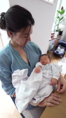 Anne, huzur içinde uyuyan, beyaz bir battaniyeye sarılı bebeğine sevgiyle bakar. Bir yuva ortamında şefkatli bir anne bakımı ve şefkat anı yakalar.