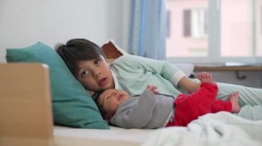 Büyük kardeş ve bebek bir yatakta uzanıyor, sıcak, güneşli bir odada, sessiz bir yakınlık ve sevgi anı paylaşıyorlar, güçlü kardeş ilişkisinin altını çiziyorlar.