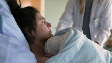 Yeni anne, doğumdan sonra acıyla boğuşuyor. Yatağa uzanmış, tıbbi ekip tarafından bakılıyor. Yeni doğmuş bebeğini göğsüne yakın tutarken battaniyelerini örtüyor.