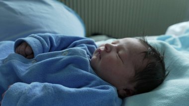 Yeni doğan bebek hastane yatağında huzur içinde uyuyor. Yumuşak mavi bir tulum giyiyor. Bebekliğinde huzurlu ve hassas bir an.