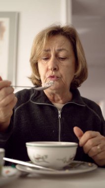 Yaşlı bir kadın yemekten zevk alıyor, her lokmanın tadını odaklanmış bir ifadeyle çıkarıyor, bir kase yiyecekle masaya oturuyor, günlük yaşamın önemini ve memnuniyetini temsil ediyor.