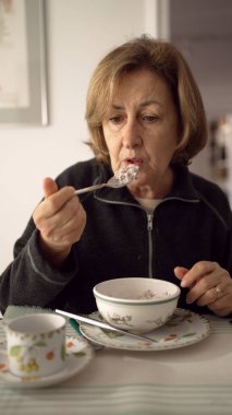 Yaşlı bir kadın, aynı tabak ve çay tabağı ile yemek yiyor, siyah ceket giyiyor, ev yemeği ortamında yemeğin tadını çıkarıyor, sağlıklı beslenme alışkanlıkları gösteriyor.