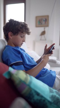 Mavi gömlekli genç, beyaz bir koltukta oturmuş akıllı telefonuna bakıyor. Çocukların ev ortamında teknolojinin modern kullanımını vurguluyor. Dijital etkileşimi vurguluyor.