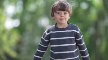 Çizgili kazaklı küçük çocuk açık havada güneşli bir günde kameraya doğru gülümsüyor.