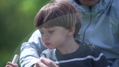 Küçük çocuk annesinin kucağında çilek yiyor parkın bahçesinde güneşin altında oturuyor.