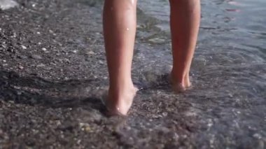 Kristal berrak gölün kıyısındaki Erkek Ayakları, Yumuşak Dalgalar Yumuşak Çakıl Zeminini okşuyor, Sıcak Yaz Güneşi Altındaki Bacakları Parıldatıyor