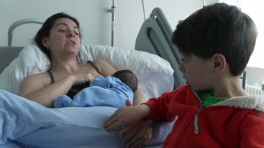 Anne hastane yatağında emzirirken, büyük kardeş yeni doğana dokunuyor. Anne dinleniyor ve kırmızı ceketli çocuk merak ve nazik bir etkileşim gösteriyor. Tıbbi ekipmanlarla hastane ortamı