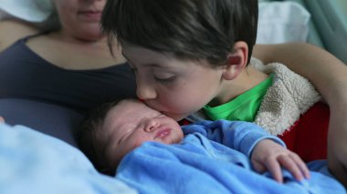 Büyük kardeş, uyuyan yeni doğmuş bebeği nazikçe öper. Annesiyle kucaklaşır. Ailevi sevgi ve bakım bağlarını vurgulayan şefkatli ve sevgi dolu bir aile anını yakalar.