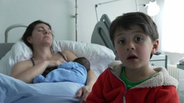 Anne hastane yatağında emzirirken, büyük kardeş yeni doğana dokunuyor. Anne dinleniyor ve kırmızı ceketli çocuk merak ve nazik bir etkileşim gösteriyor. Tıbbi ekipmanlarla hastane ortamı