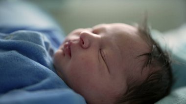 Serene bebek mavi bir tulumla uyuyor, hastane yatağında yumuşak battaniyelerle kundaklanmış, bebeğin huzurlu ifadesine odaklanıyor, yeni doğan hayatının sükunetini yakalıyor.