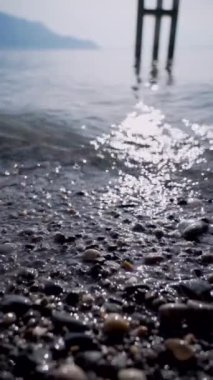 Güneş, berrak bir gölün kenarında, arka planda dağlar ve görülebilir kıyı taşları olan suya yansıyor.